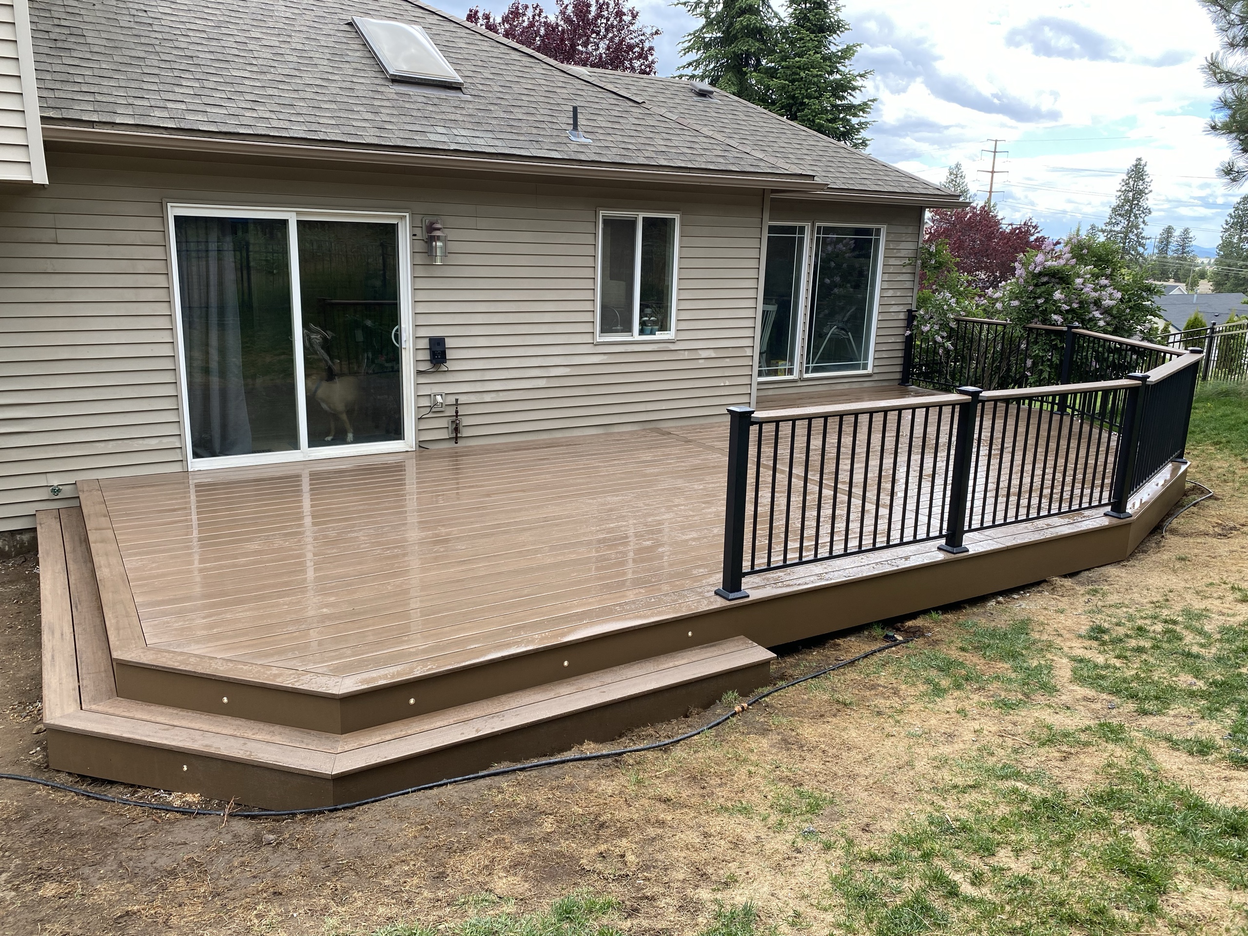 Wet brown composite deck with step lights and black picket railing at back of tan house, dry lawn