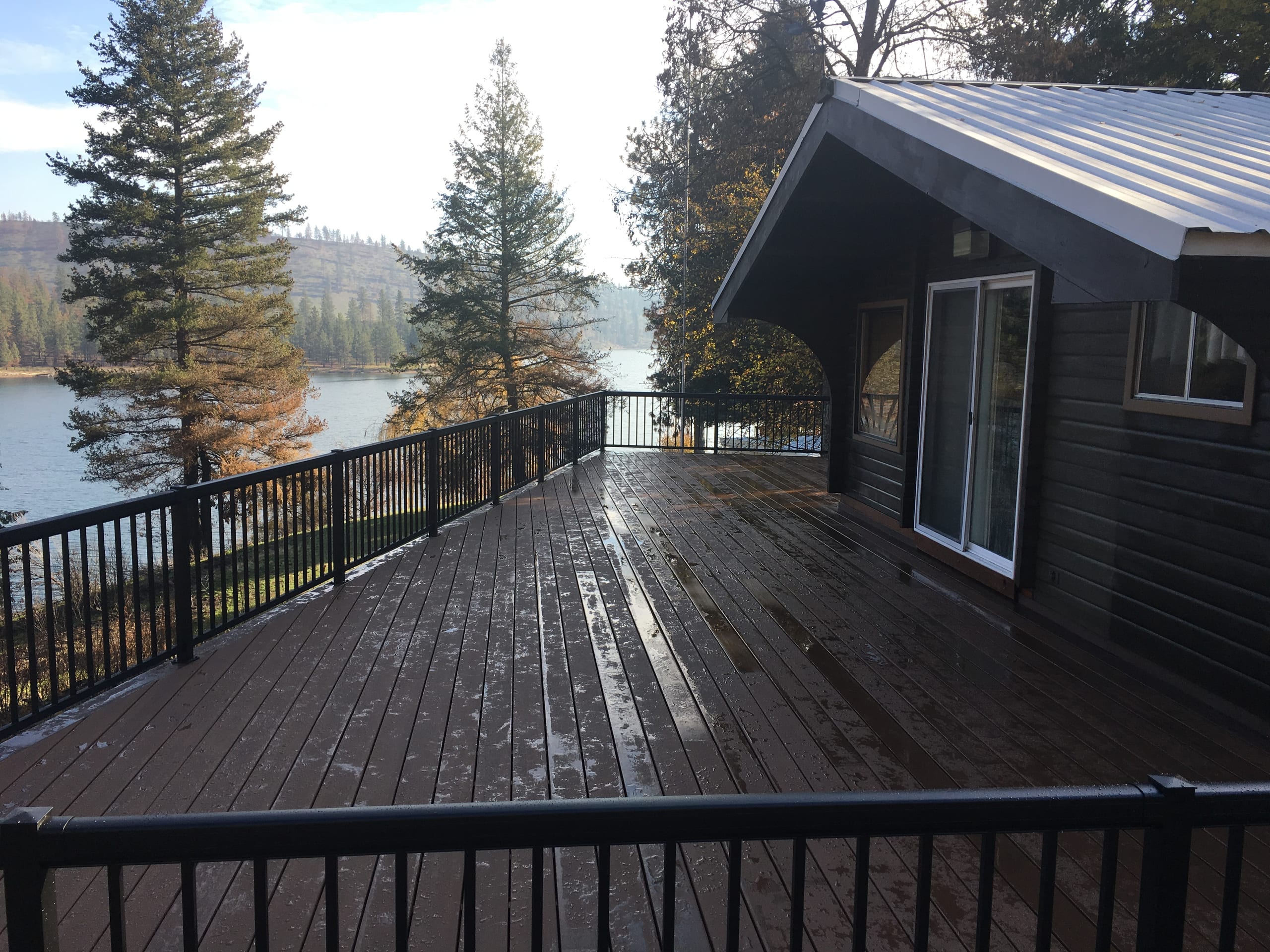 Wet dark brown deck with black picket railing outside a dark-sided house on a lake, pines and metal roof