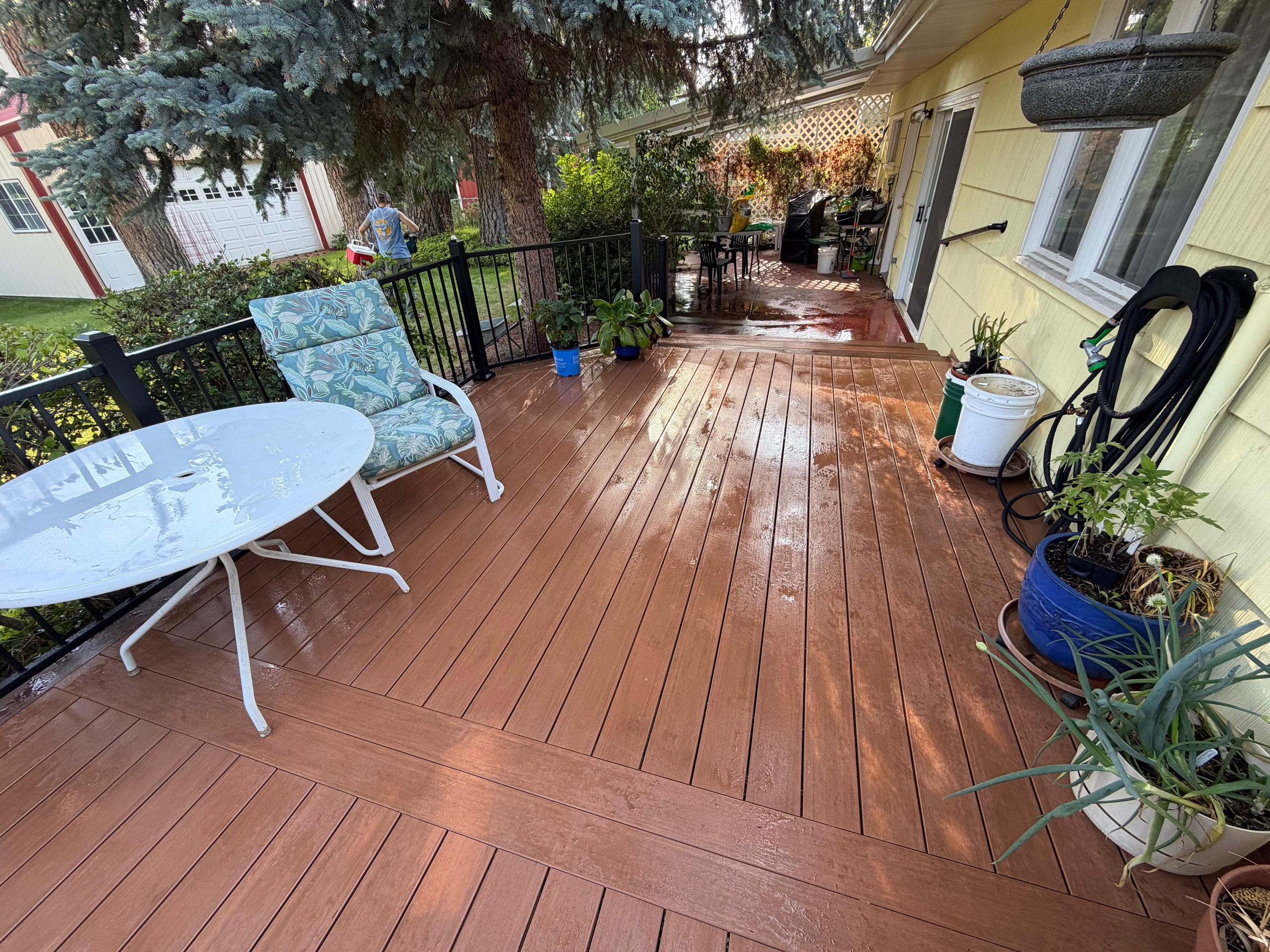Wet reddish-brown composite deck with black metal railing, patio table, chairs and potted plants beside yellow house