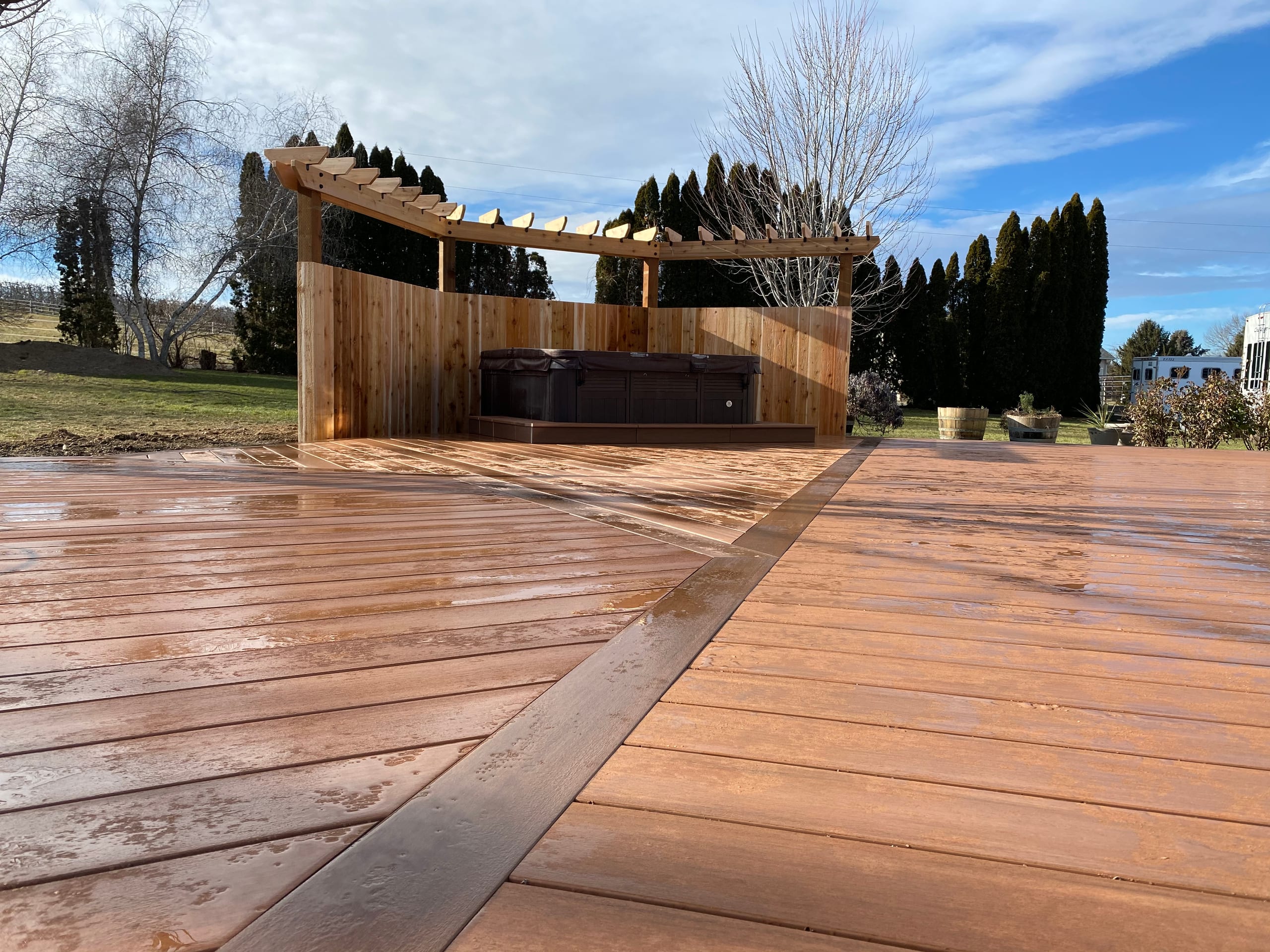 Wet reddish composite deck with border inlay and cedar privacy wall pergola around hot tub, open fields beyond