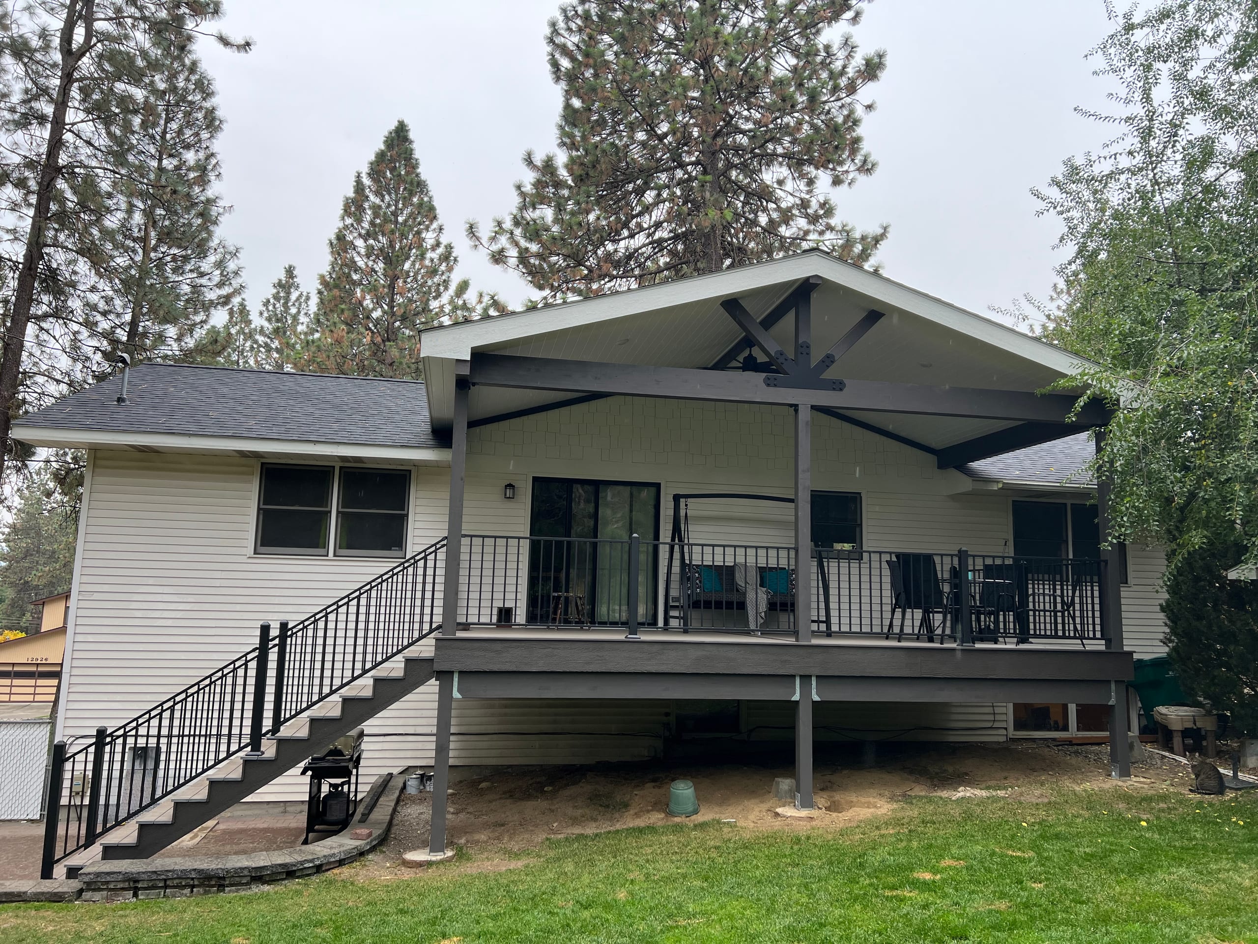 White house with new elevated covered deck, dark gabled roof with trusses, black railing and stair, bark and lawn below