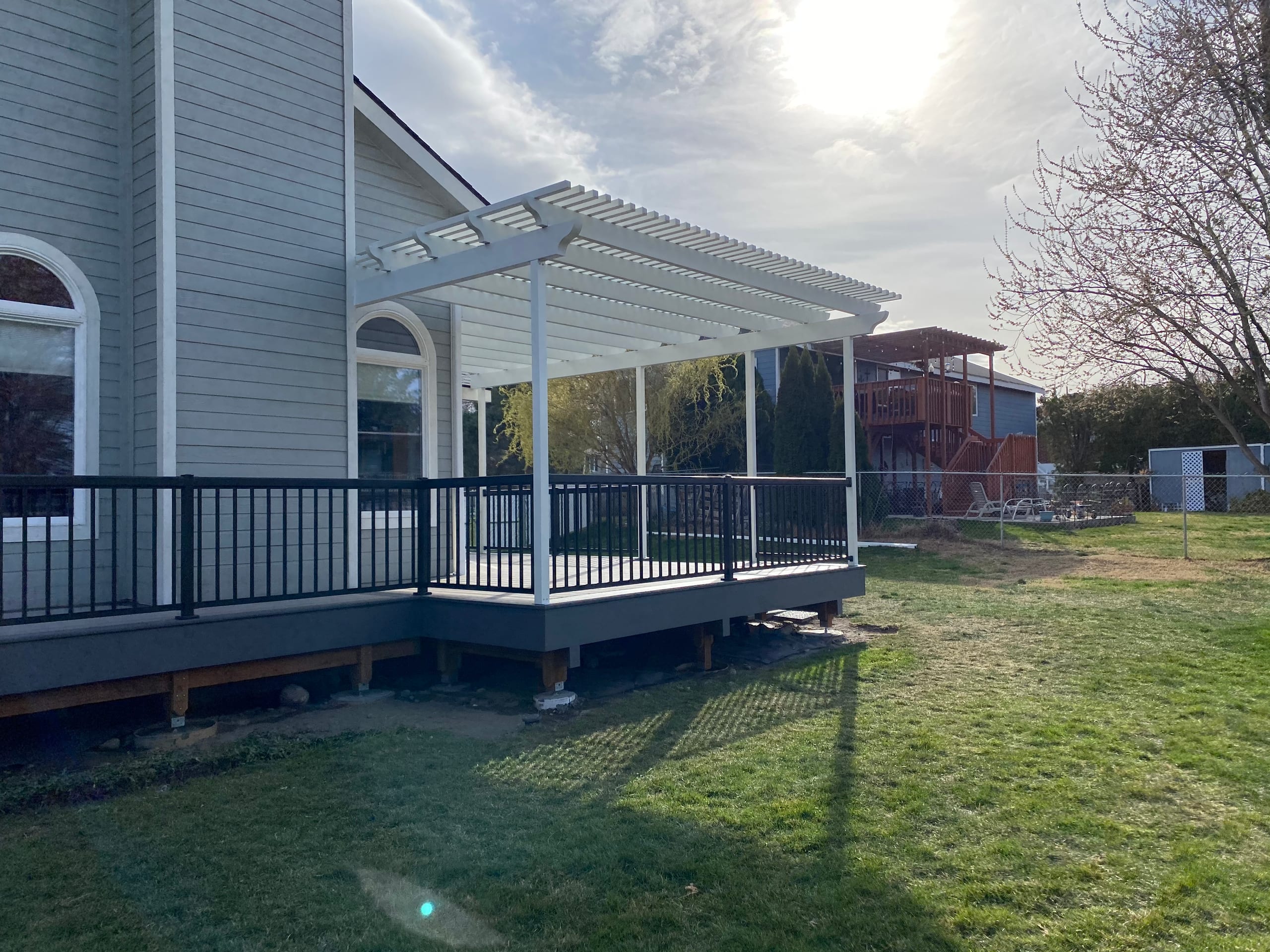 White louvered pergola over a grey composite deck with black metal railing beside a tan house with large lawn