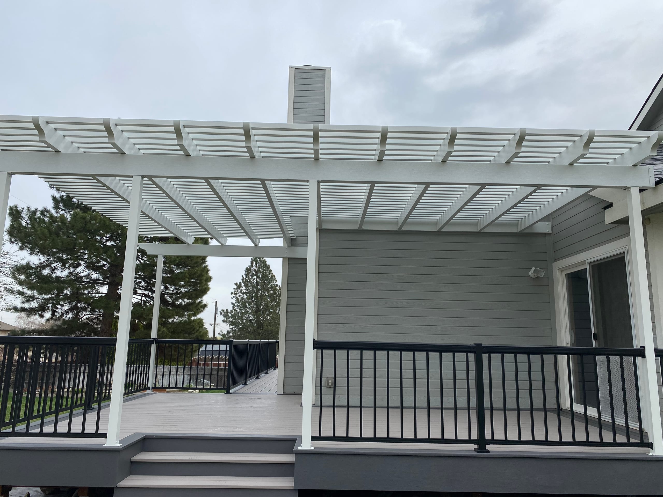 Under the white louvered pergola on a light gray composite deck with black picket railing and steps