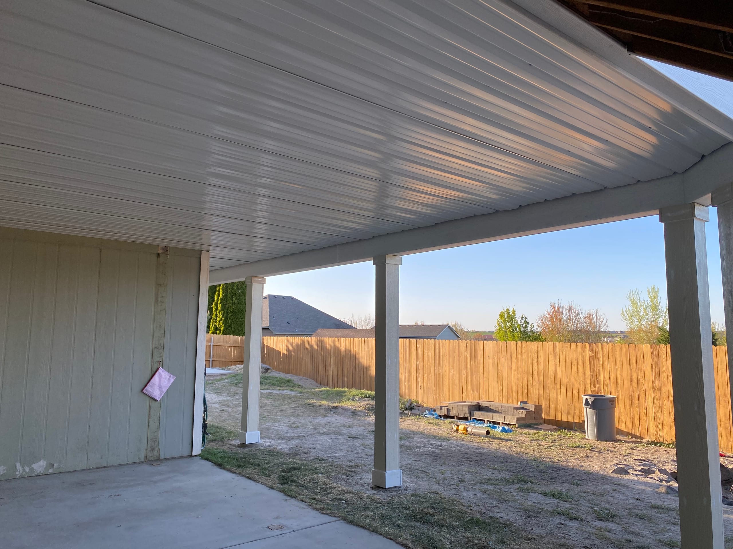 White underdeck ceiling and white posts over a concrete patio, backyard fence and some materials beyond