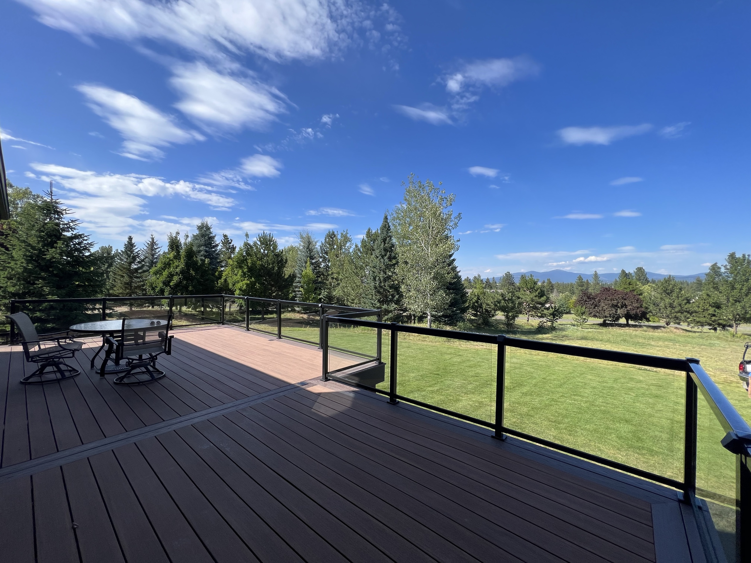 Wide brown composite deck with glass-panel railing, patio furniture, lawn, tree line and blue sky
