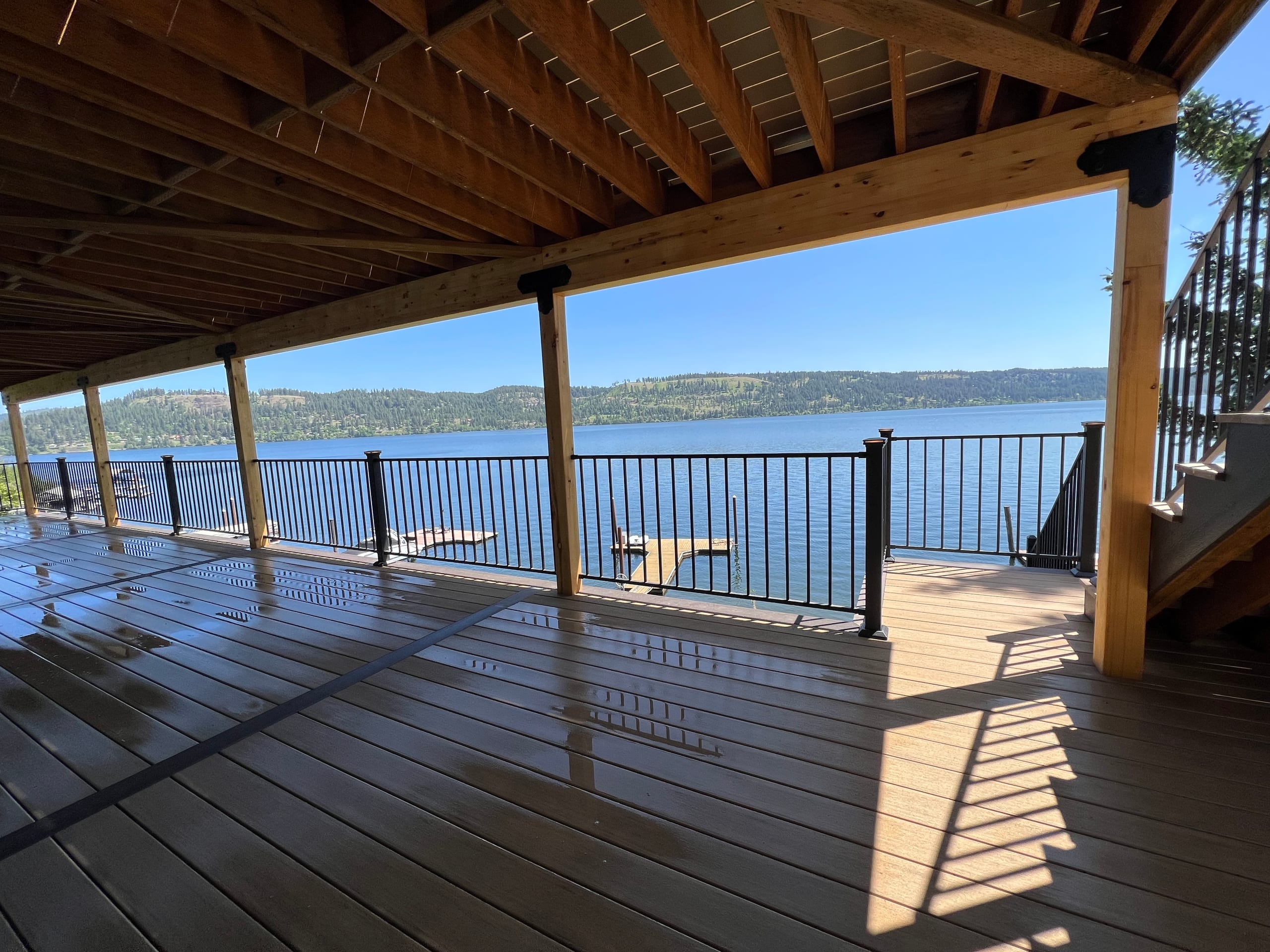 Wide view of covered lakeside deck with exposed joist ceiling, black railing, posts and stairs, lake beyond