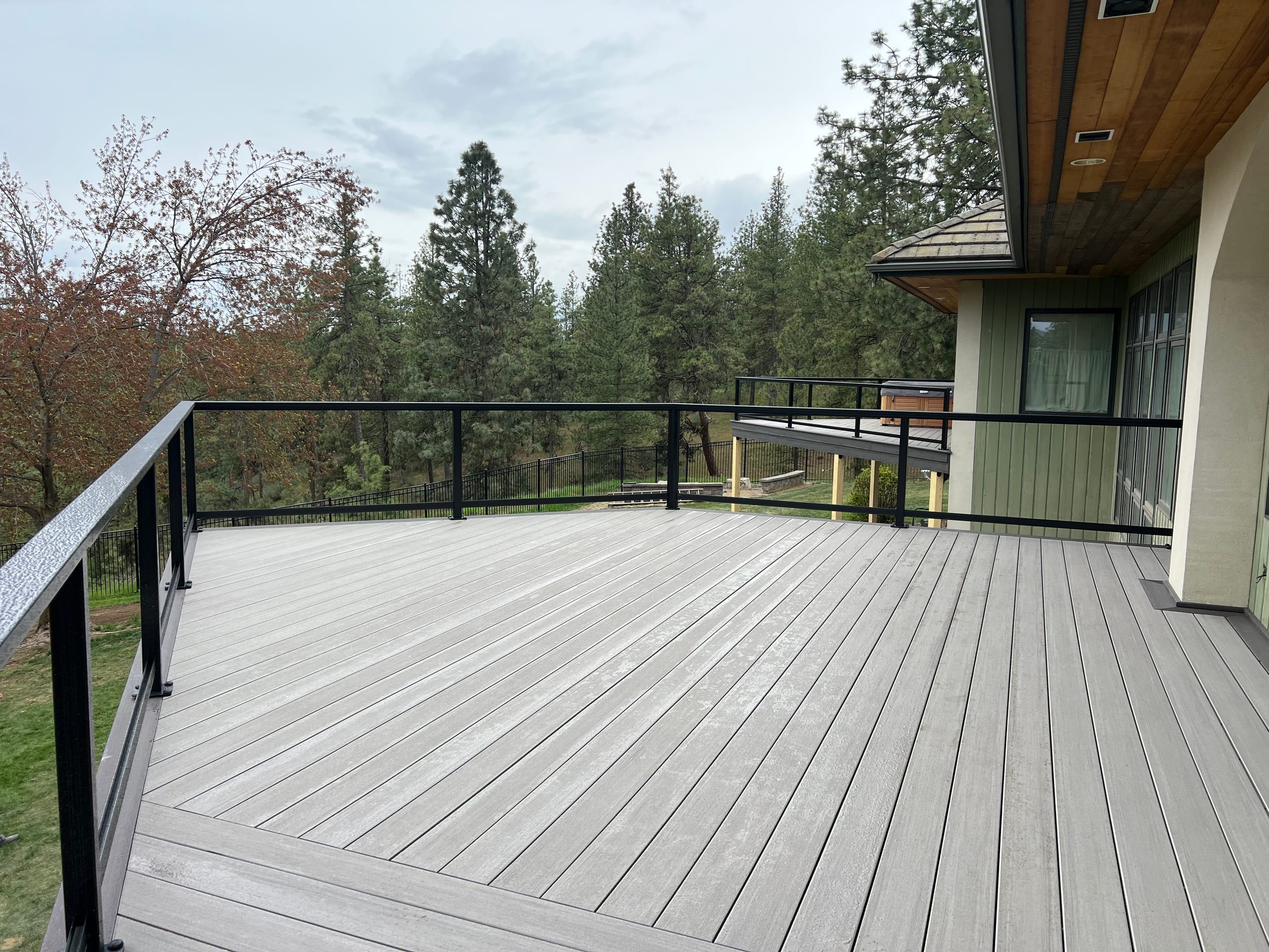 Wide light gray composite deck with black railing, wood soffit and pine forest views, overcast sky