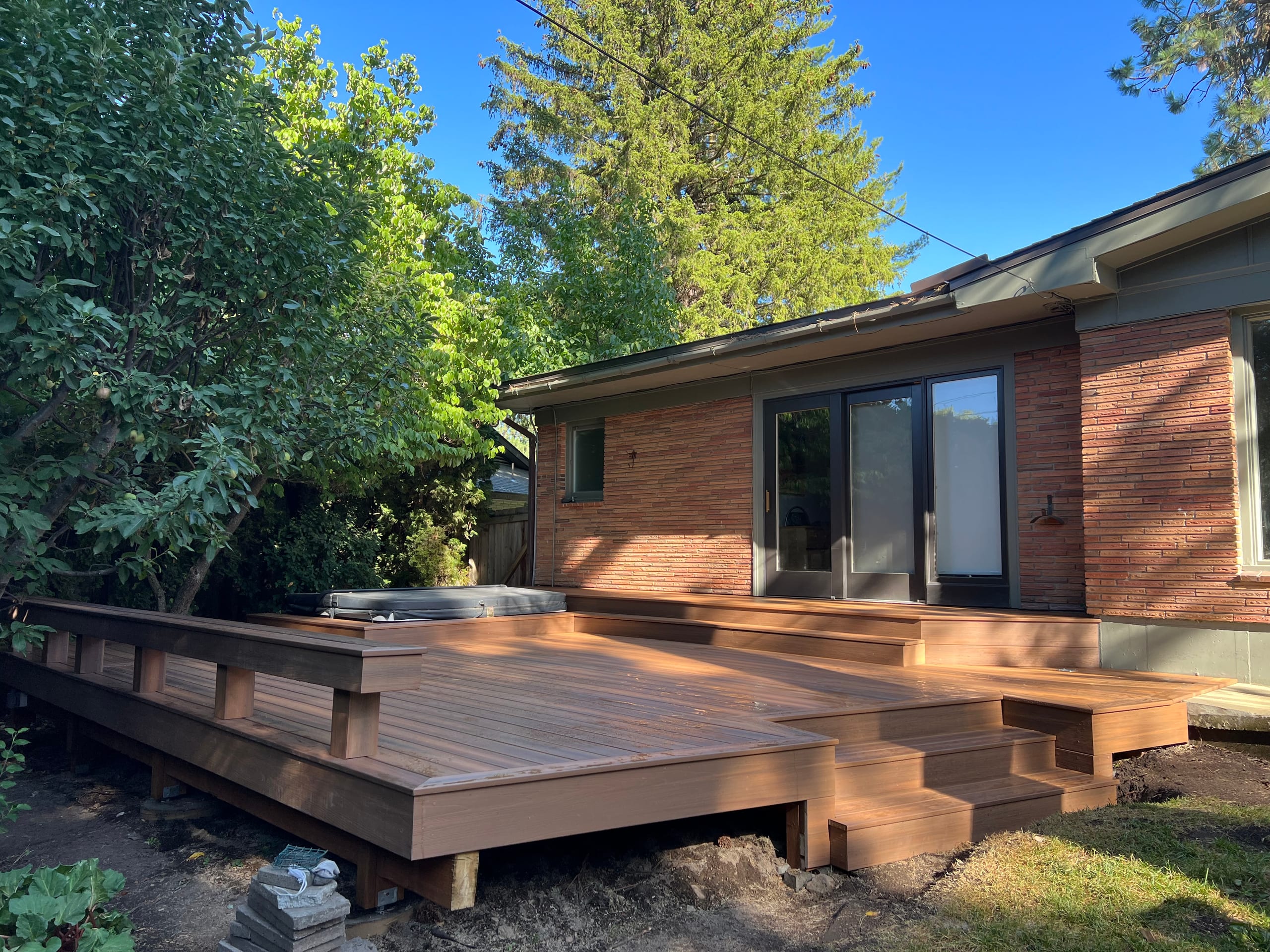 Wide low red-brown deck with steps, perimeter bench and hot tub behind a brick ranch house