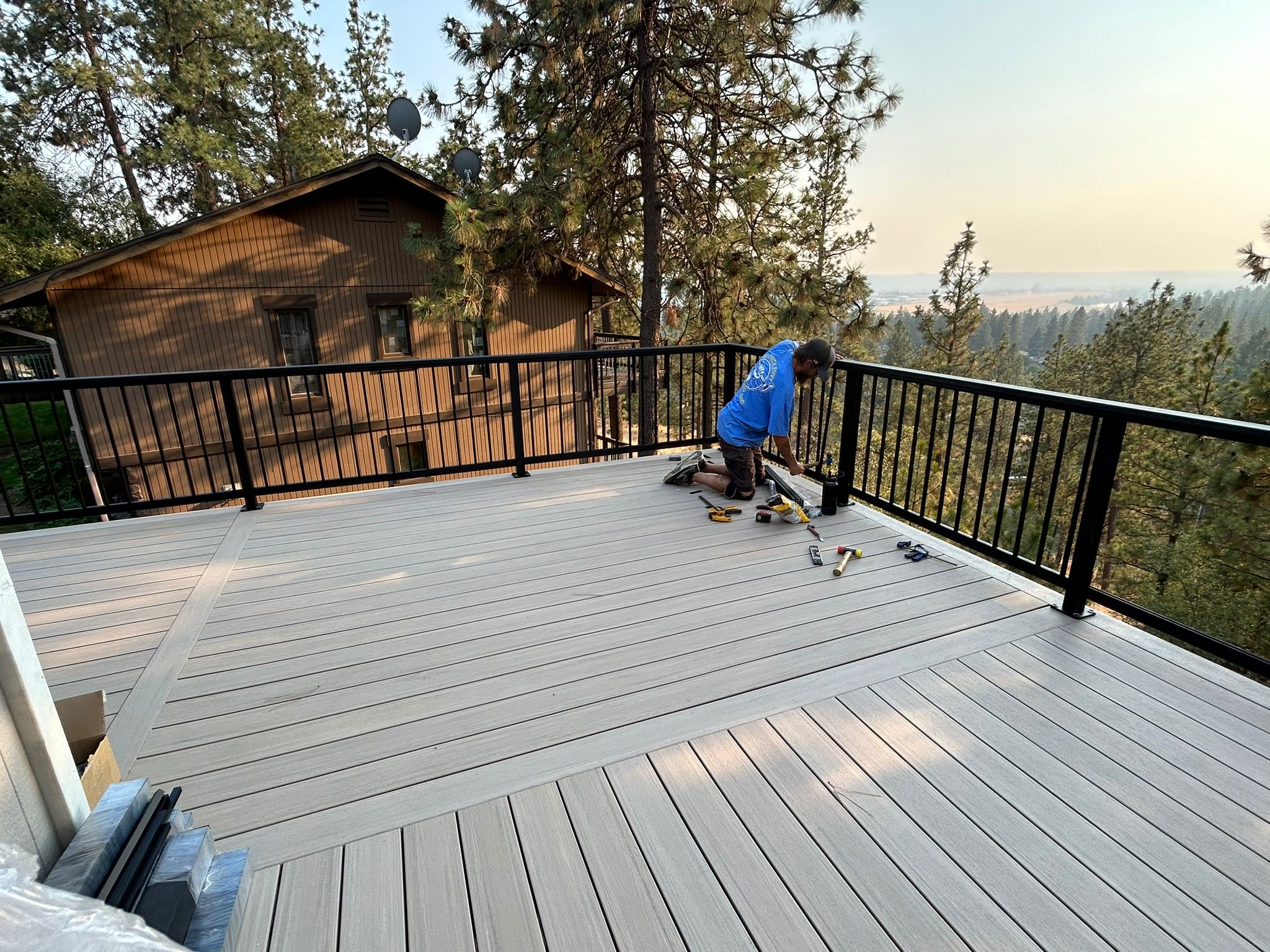 Worker installing light gray composite decking on an elevated deck with black railing and a hillside view at golden hour